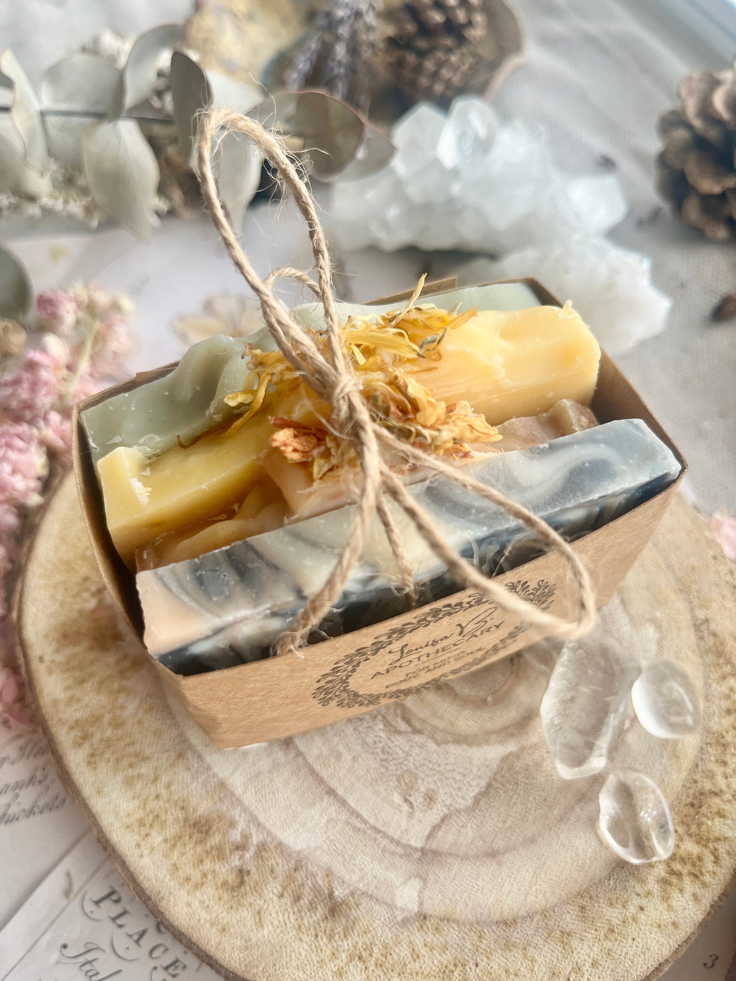 Four slices of soap in decorative packaging with twine on a wooden surface with dried flowers , eucalyptus and crystals