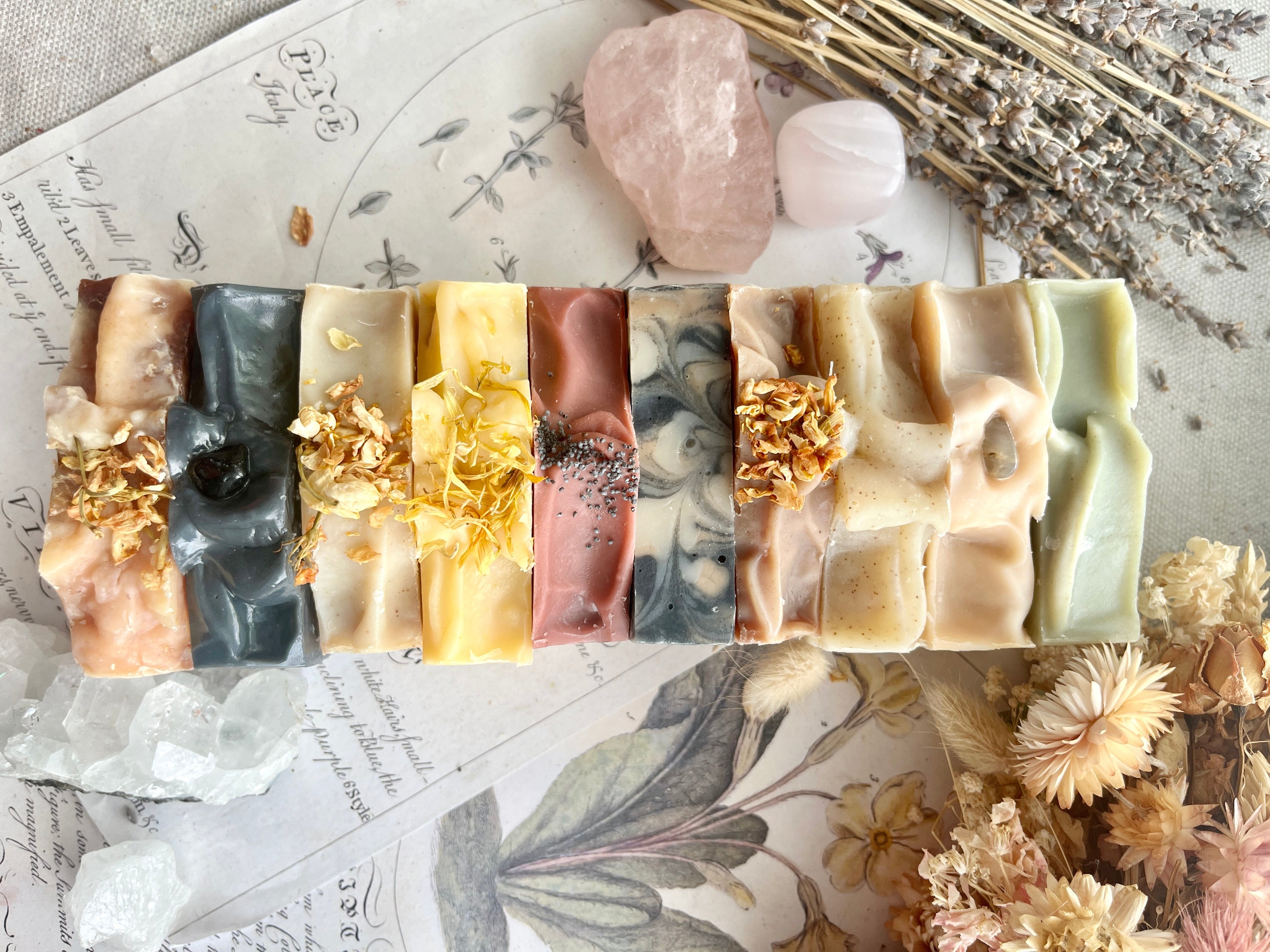 A vertical stack of ten artisanal handmade soap bars in various colors and patterns, decorated with dried flowers and botanicals, set on a vintage-style botanical print background with crystals.