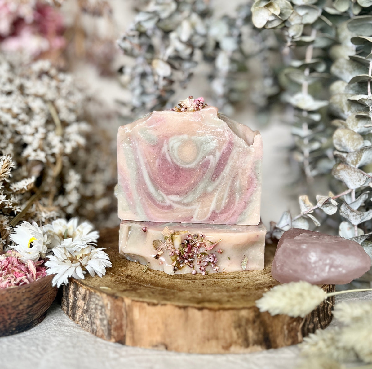 Two handmade Louisa B Apothecary soap bars stacked on a rustic wood slice, decorated with dried flowers, surrounded by eucalyptus leaves, white daisies, and a rose quartz crystal.