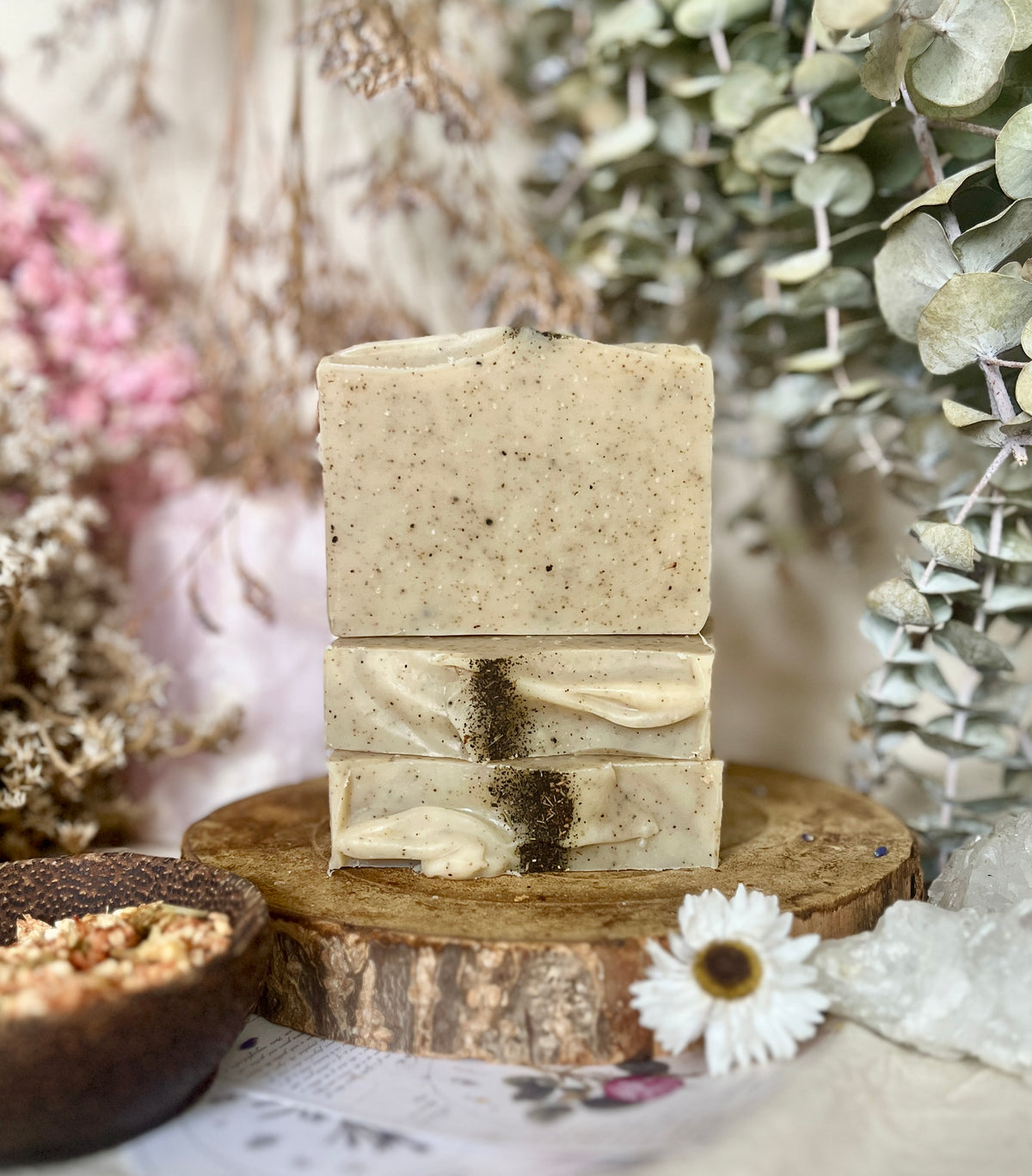 Three stacked bars of Louisa B Apothecary handmade soap on a rustic wood slice, surrounded by dried flowers, greenery, and a small bowl in a styled natural setting.