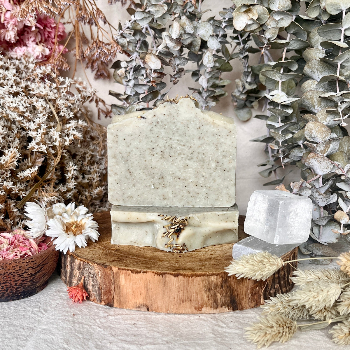 A Louisa B Apothecary handmade soap bar with a rustic, textured light green surface sits on a wooden slice pedestal, surrounded by dried flowers, eucalyptus leaves, and crystals.