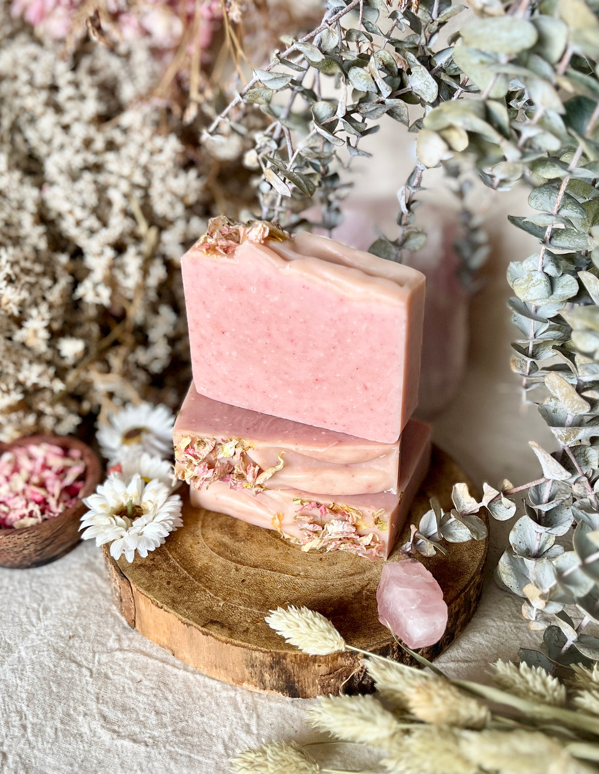 Two stacked pink handmade Louisa B Apothecary soap bars with dried flower petals, displayed on a rustic wooden slice surrounded by dried flowers, eucalyptus, and a pink rose quartz crystal.
