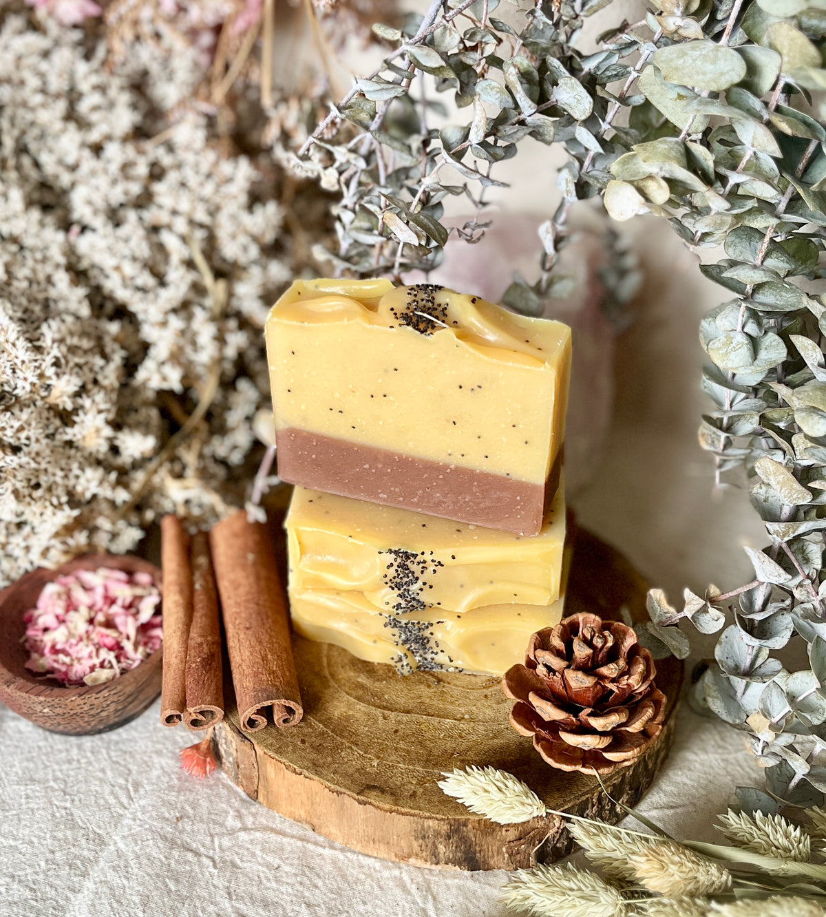 Two stacked handmade soap bars with a yellow and brown color-blocked design, decorated with black seeds, displayed on a rustic wooden slice surrounded by dried flowers, cinnamon sticks, and a pinecone.