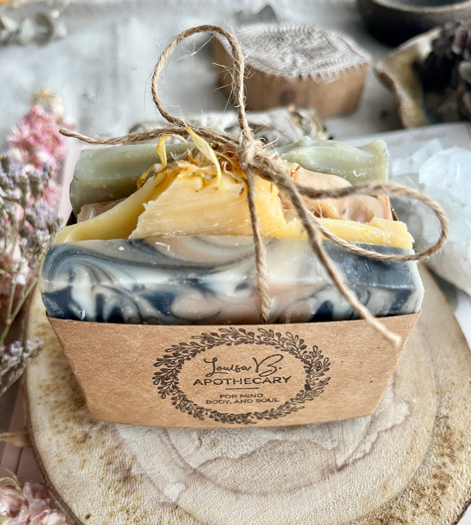 Four soaps in a packaged with twine, branded 'Louisa B& Apothecary', on a wooden surface, surrounded by botanicals