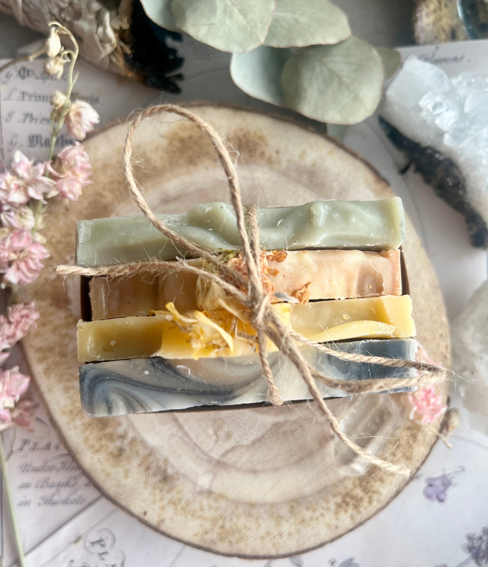 Four bars of soap stacked on a round wooden surface with twine, surrounded by flowers and leaves.