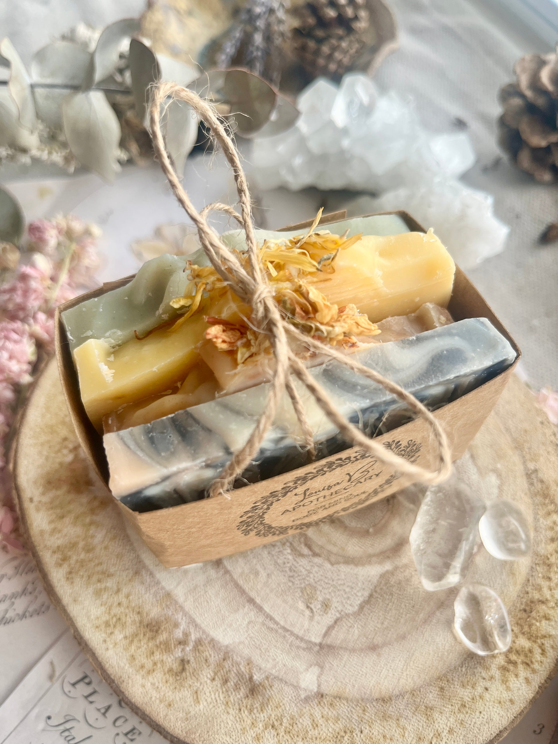 Four slices of soap in decorative packaging with twine on a wooden surface with dried flowers , eucalyptus and crystals