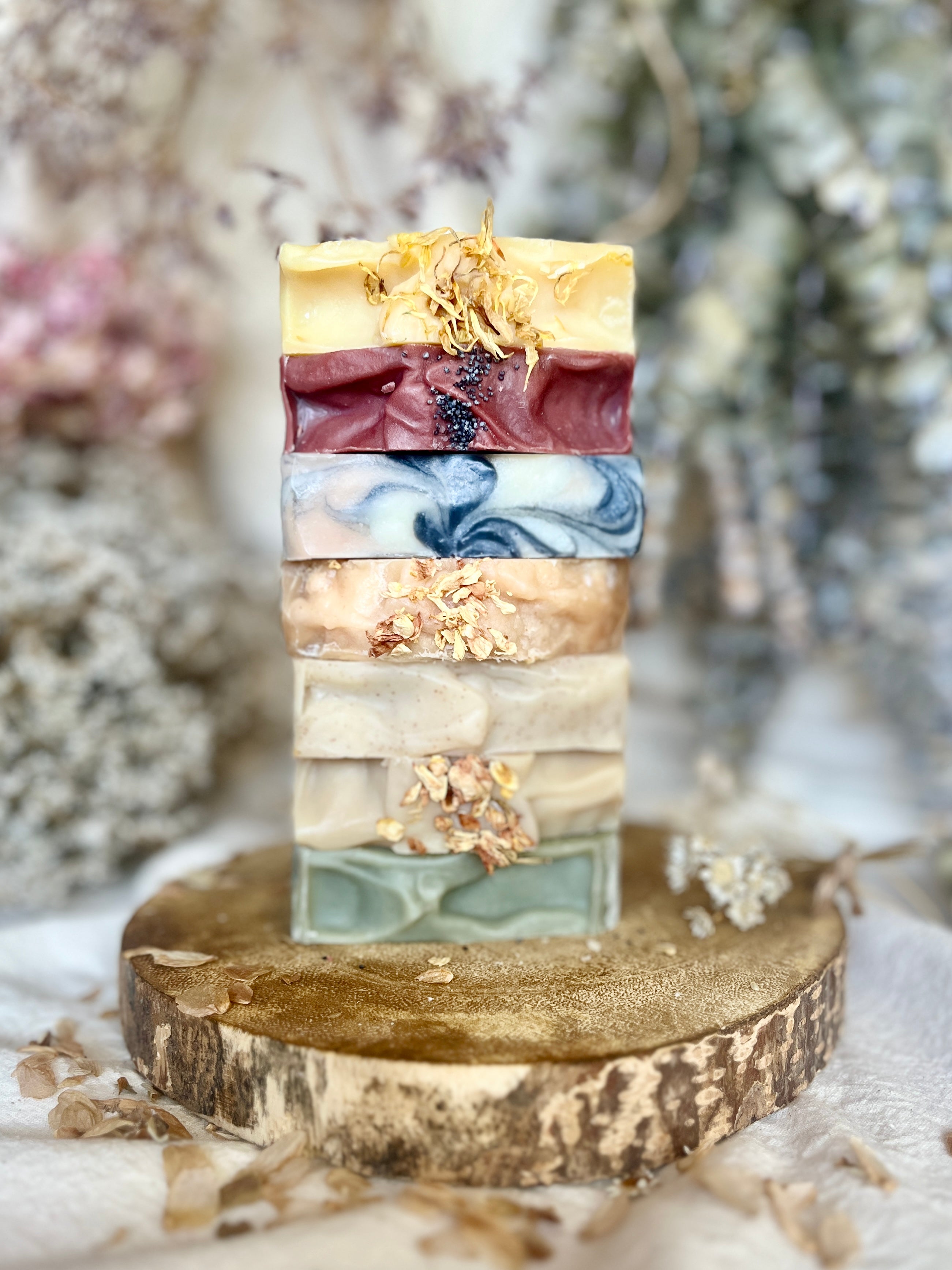 A stack of six artisanal handmade soap bars in various earthy colors, each topped with dried flower petals, displayed on a rustic wood slice stand.