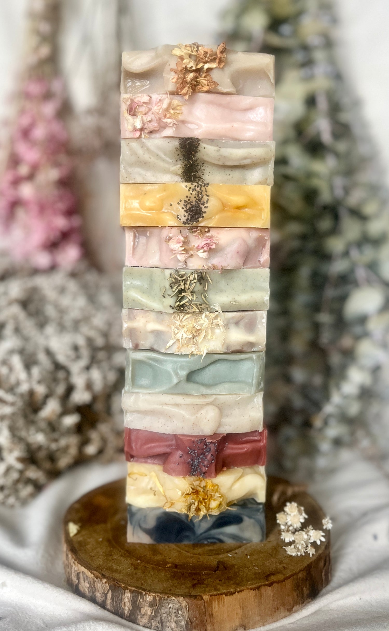 A vertical stack of ten artisanal handmade soap bars in various pastel and earthy colors, each decorated with dried botanicals, displayed on a rustic wooden slice coaster against a soft, blurred background of dried flowers.